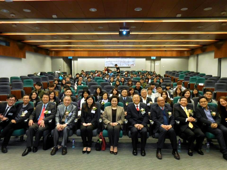 A lecture hall full of seated people facing the camera; the people in the front row wear suits and corsages.