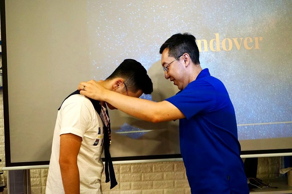 A person in a blue polo shirt places a ceremonial collar over the bowed head of a person in a white T-shirt, in front of a projection screen.