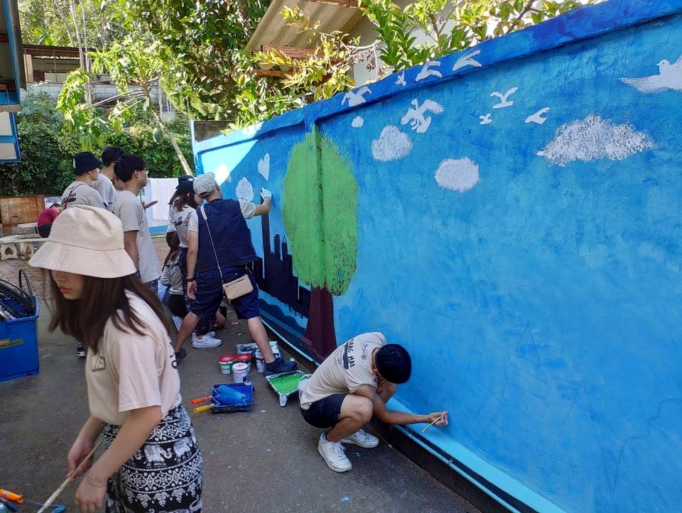 Several people paint a mural on a long wall: a blue sky with white clouds and birds, a green tree and a dark skyline; one crouches to paint the base of the wall.