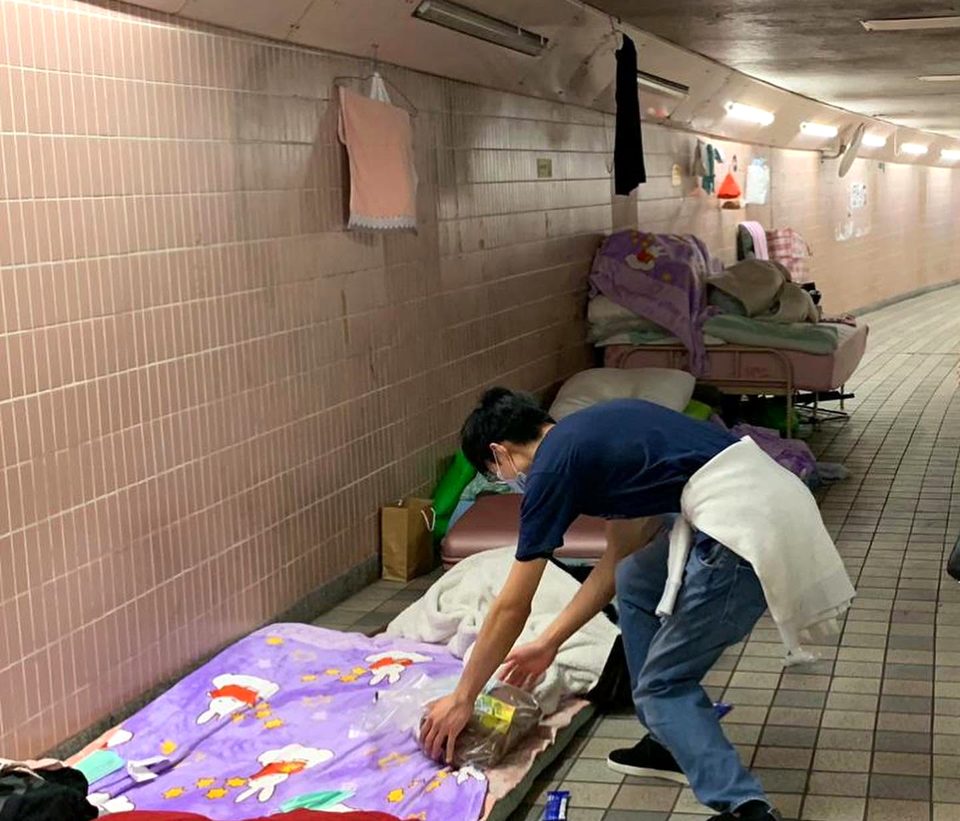 In a tiled pedestrian subway, a person in a face mask bends down to set a jar on bedding laid out along the wall.