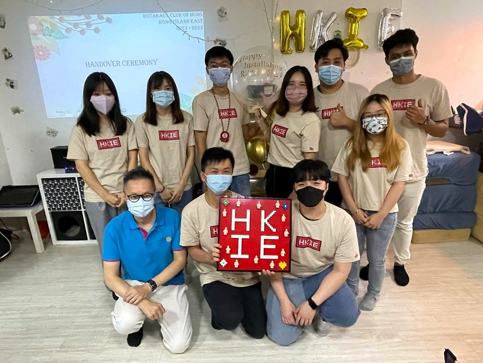 A group of people in face masks, most in matching beige T-shirts printed HKIE, pose in two rows; one holds a red plaque that spells HKIE, and gold balloons hang behind.